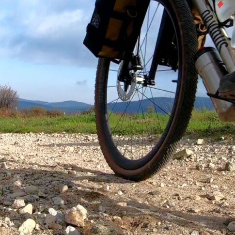 vélo titane gravel titane bikepacking obvious outquest roulant sur chemin de caillou, vu du bas. on voit les bagages akao designs et les chaussures du cycliste